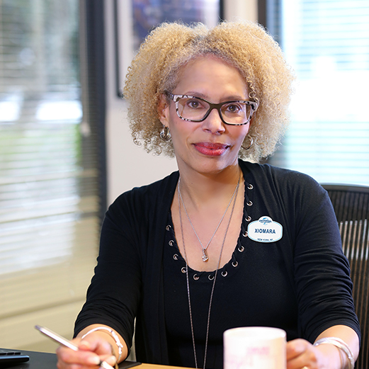 Marketing & sales team member seated a desk with a pen in one hand and mug in another.