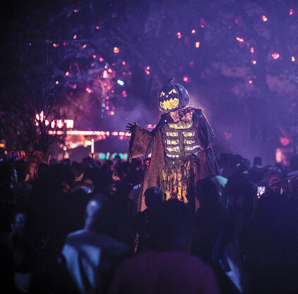 A performer on stilts dressed as a scarecrow with a pumpkin head that glows towers over guests walking around them.