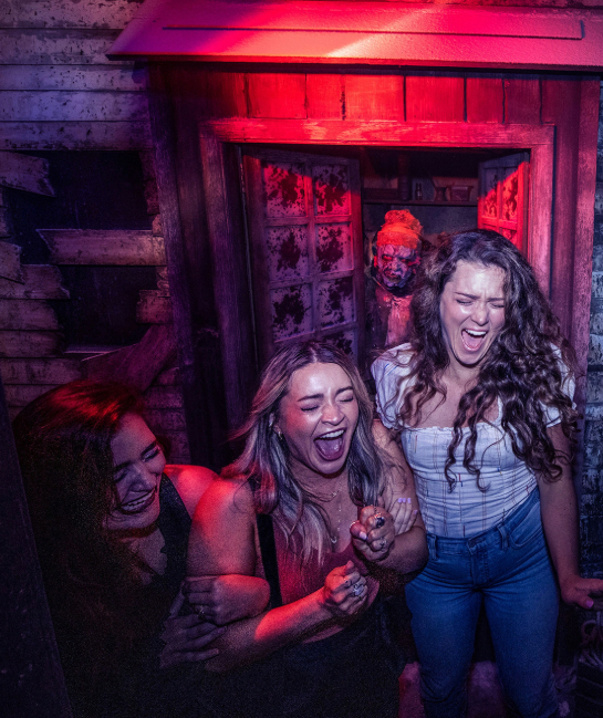 three women screaming in a haunted house, getting scared by performer behind them in a doorway.