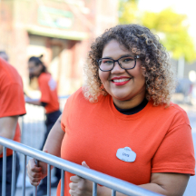 A smiling woman wearing an orange shirt stands next to a metal fence outside a haunted house.